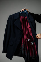 A dark navy blue blazer jacket is held open by a hand on a hanger, revealing a rich burgundy satin lining with a subtle black damask pattern. The blazer features a notch lapel, flap pockets, and a black brand patch on the pocket.