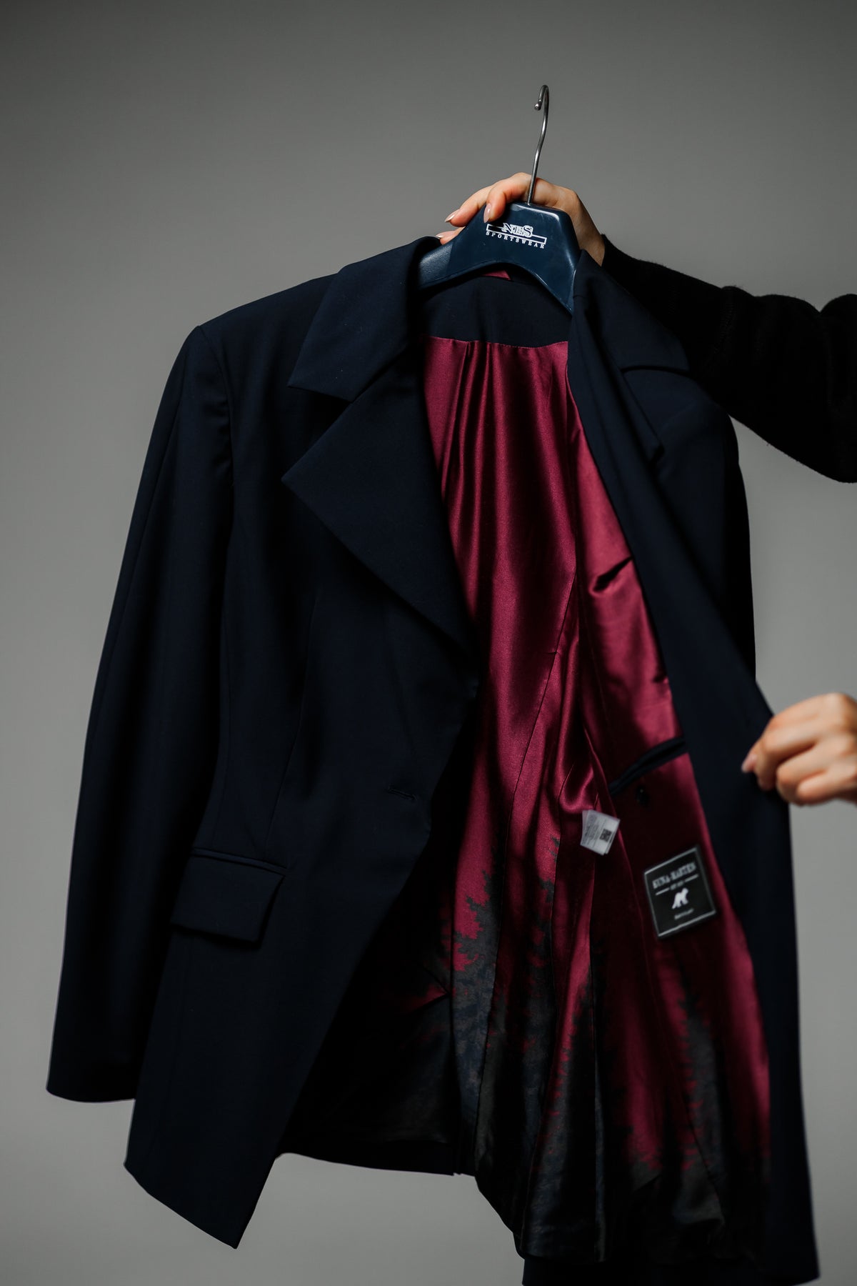 A dark navy blue blazer jacket is held open by a hand on a hanger, revealing a rich burgundy satin lining with a subtle black damask pattern. The blazer features a notch lapel, flap pockets, and a black brand patch on the pocket.