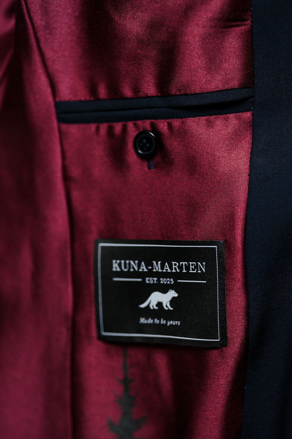 Close-up of a burgundy satin lapel with a black button and a black Kuna-Marten brand patch sewn onto the inside.