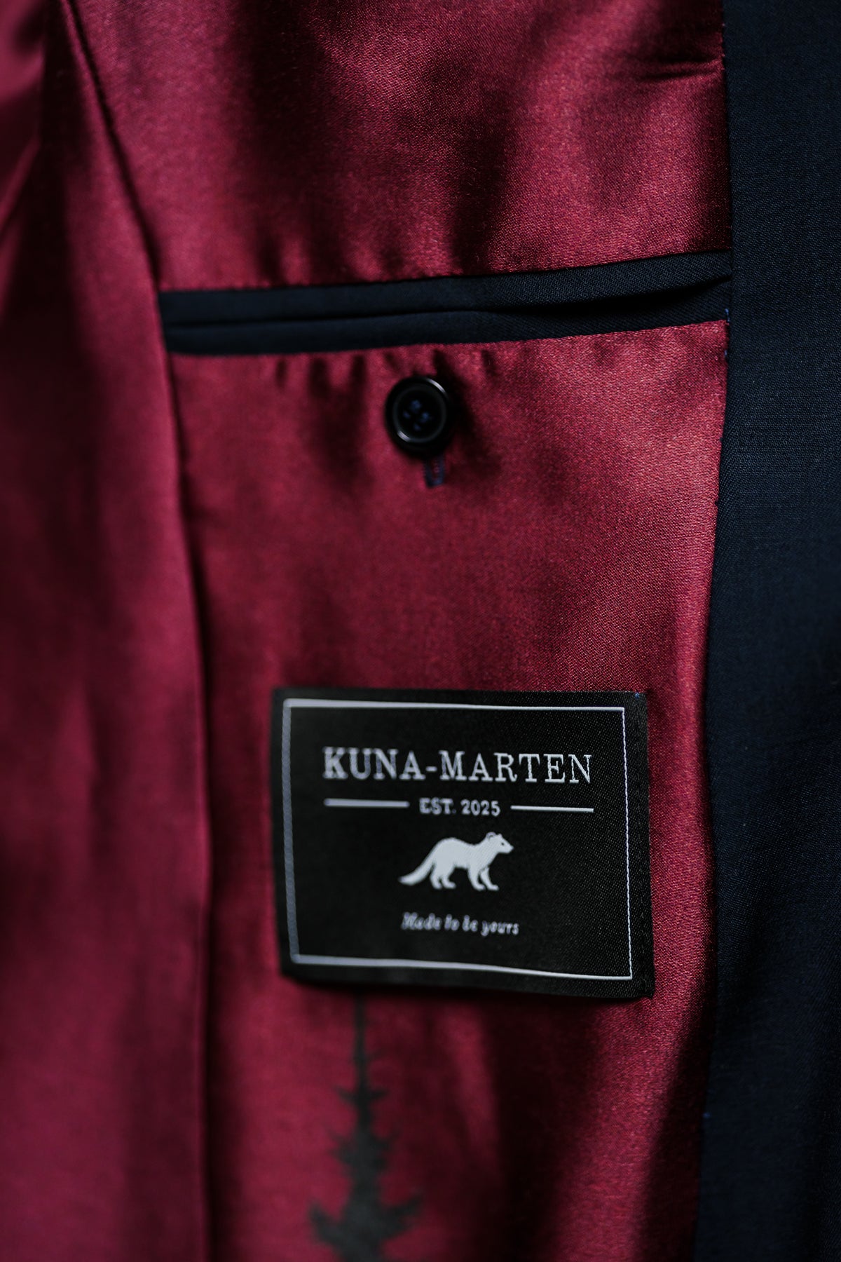Close-up of a burgundy satin lapel with a black button and a black Kuna-Marten brand patch sewn onto the inside.